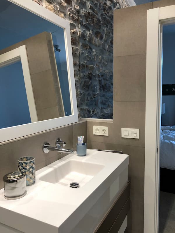 Griferia del lavabo del baño sale directamente de la pared. Espejo con marco blando. Cristalera en la parte de arriba de las paredes para que entre luz natural